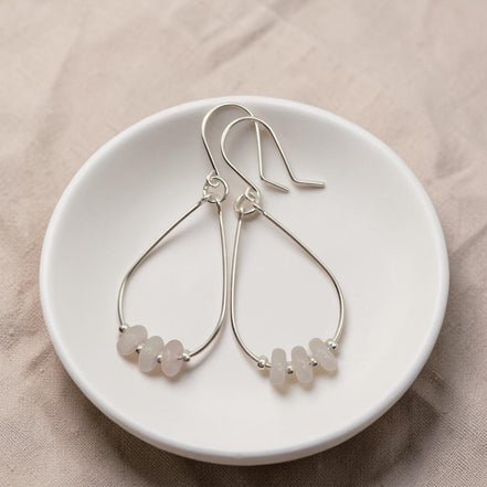 Bijou by SAM Aurora teardrop hoop earrings featuring blush rose quartz beads on sterling silver wire in a white dish.