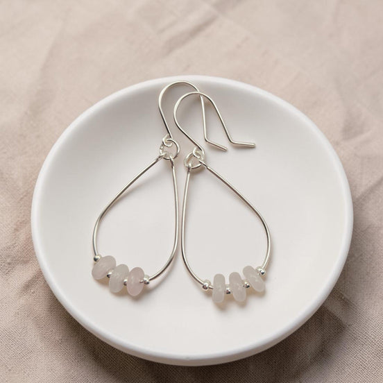 Bijou by SAM Aurora teardrop hoop earrings featuring blush rose quartz beads on sterling silver wire in a white dish.