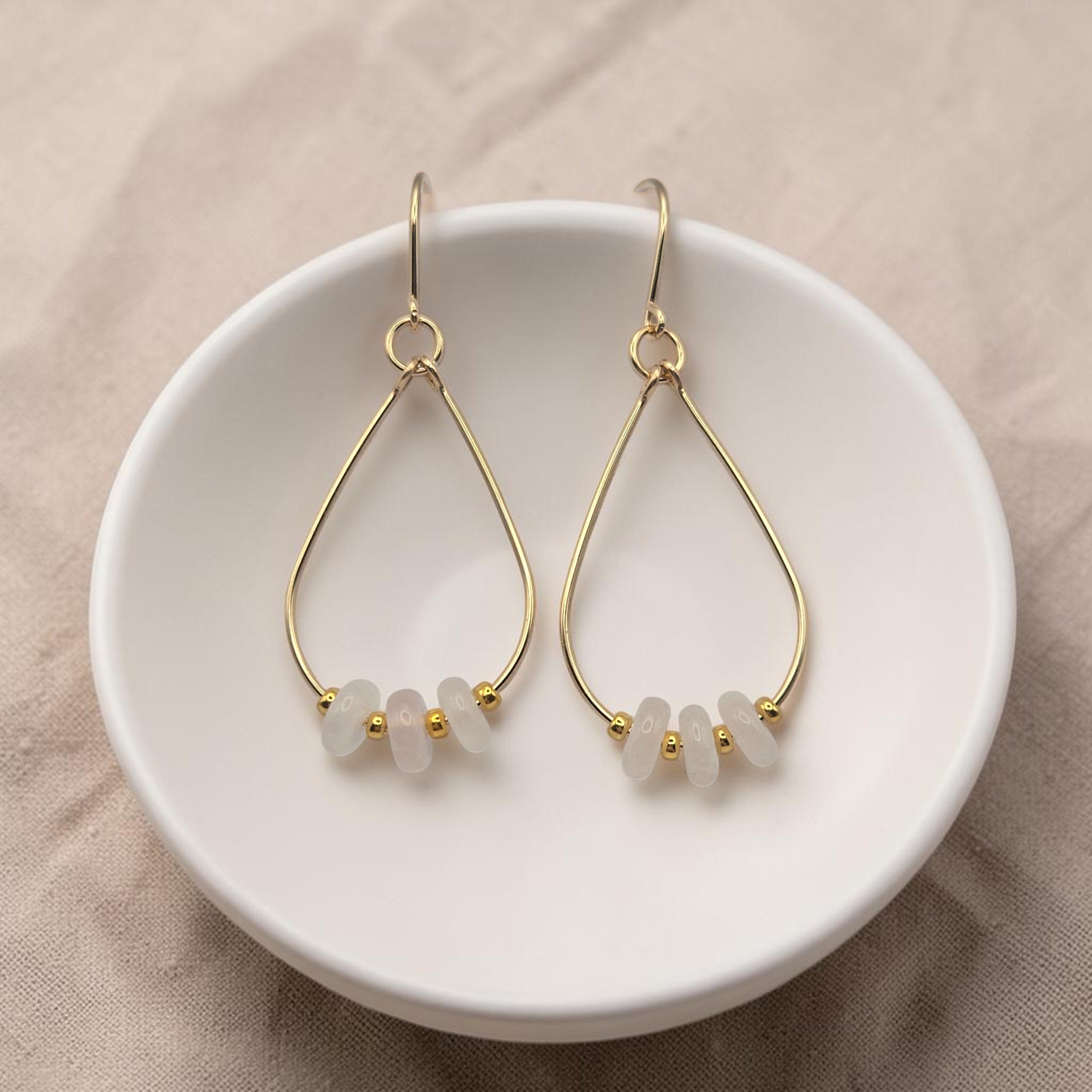 Bijou by SAM Aurora teardrop hoop earrings featuring blush rose quartz beads and 14k gold fill wire in a white dish.