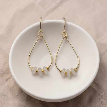 Bijou by SAM Aurora teardrop hoop earrings featuring blush rose quartz beads and 14k gold fill wire in a white dish.