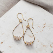 Bijou by SAM Aurora teardrop hoop earrings featuring blush rose quartz beads on 14k gold fill wire on stone surface.