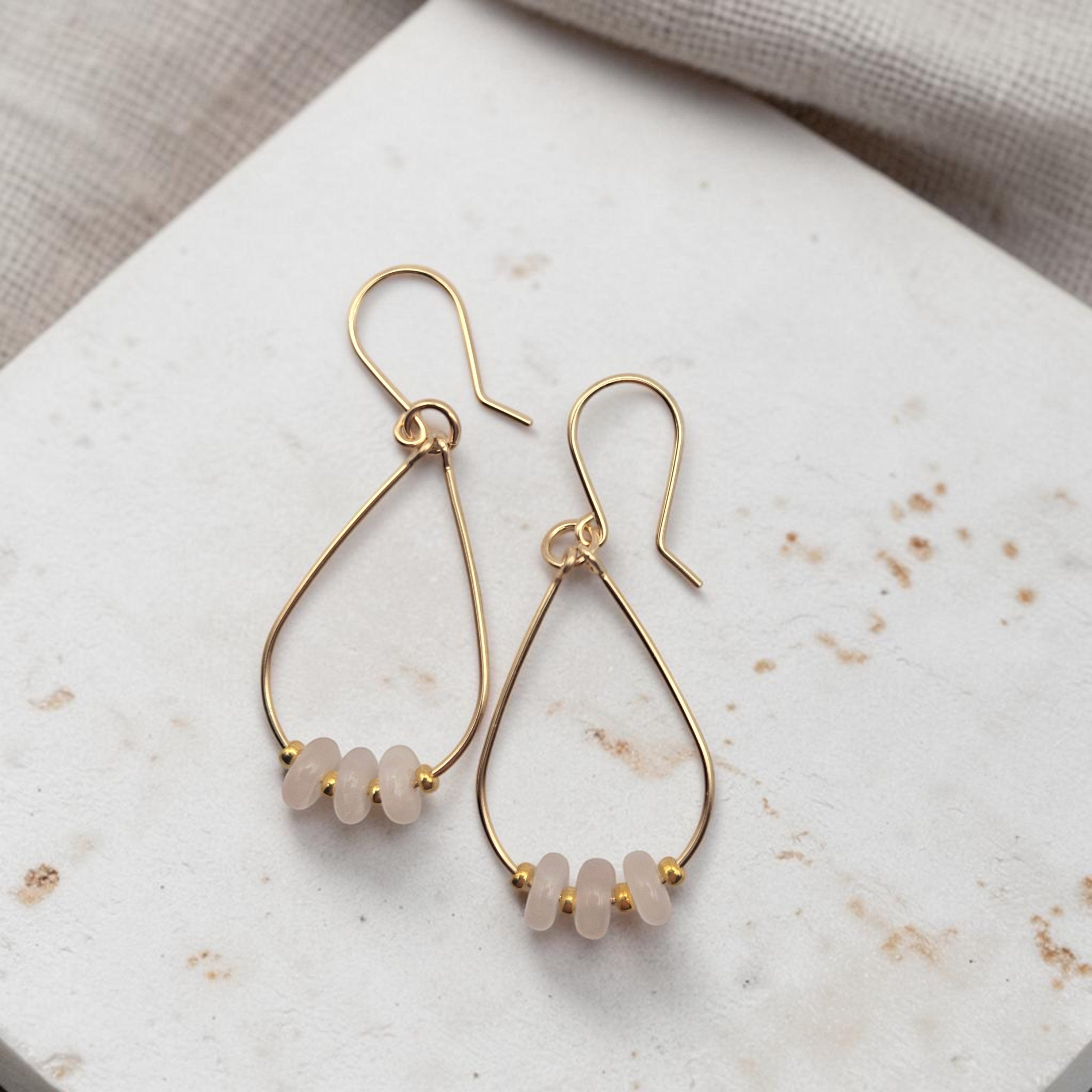 Bijou by SAM Aurora teardrop hoop earrings featuring blush rose quartz beads on 14k gold fill wire on stone surface.