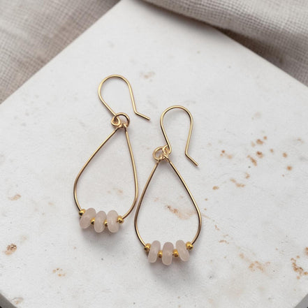 Bijou by SAM Aurora teardrop hoop earrings featuring blush rose quartz beads on 14k gold fill wire on stone surface.