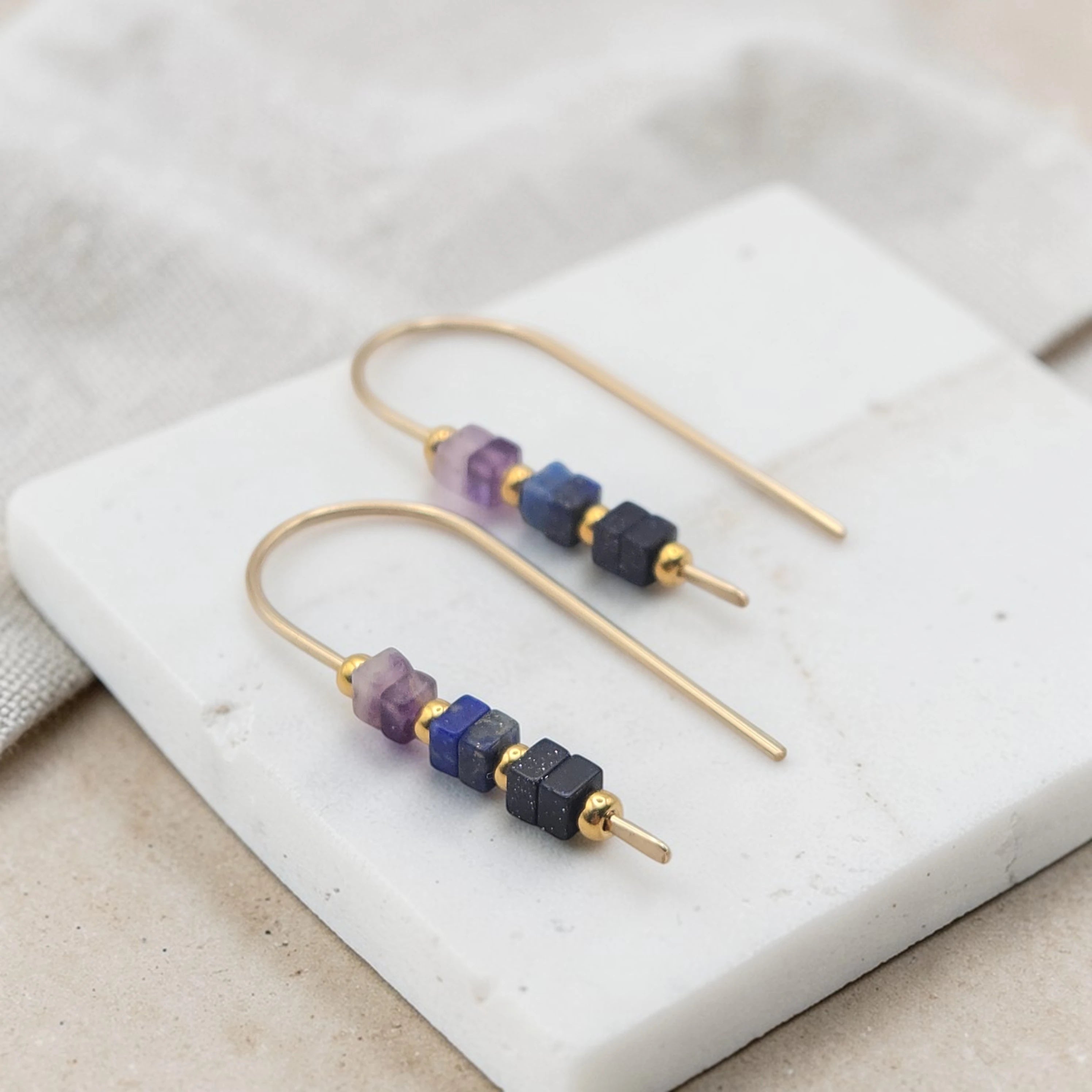 Gold earrings with purple and blue beads on a white stone surface