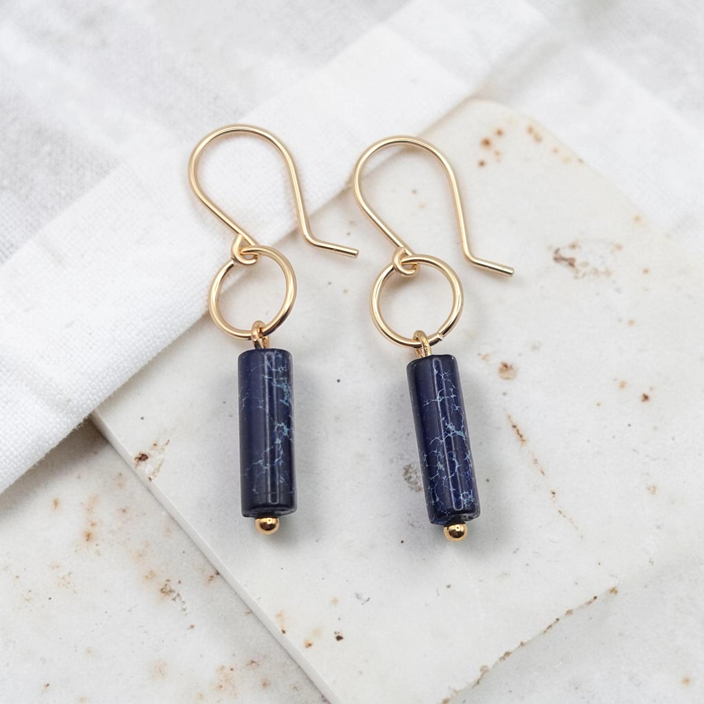 Pair of gold earrings with navy blue cylindrical beads on a marble surface.