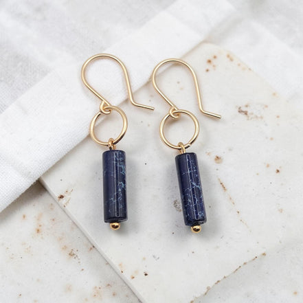 Pair of gold earrings with navy blue cylindrical beads on a marble surface.
