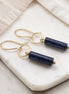 Pair of gold earrings with navy blue cylindrical stones on a marble surface.