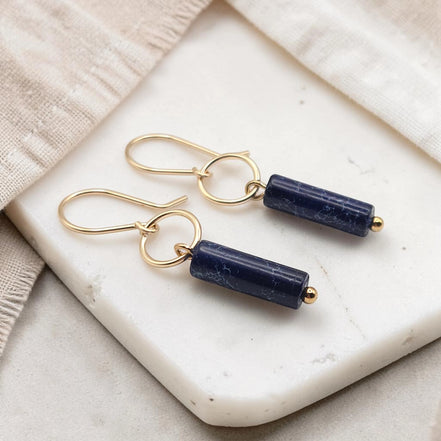 Pair of gold earrings with navy blue cylindrical stones on a marble surface.