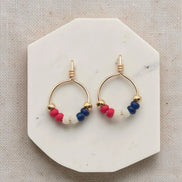 Pair of gold hoop earrings with red, blue, and white beads on a marble stand.