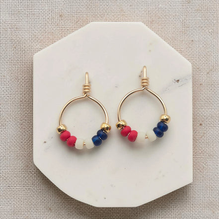 Pair of gold hoop earrings with red, blue, and white beads on a marble stand.