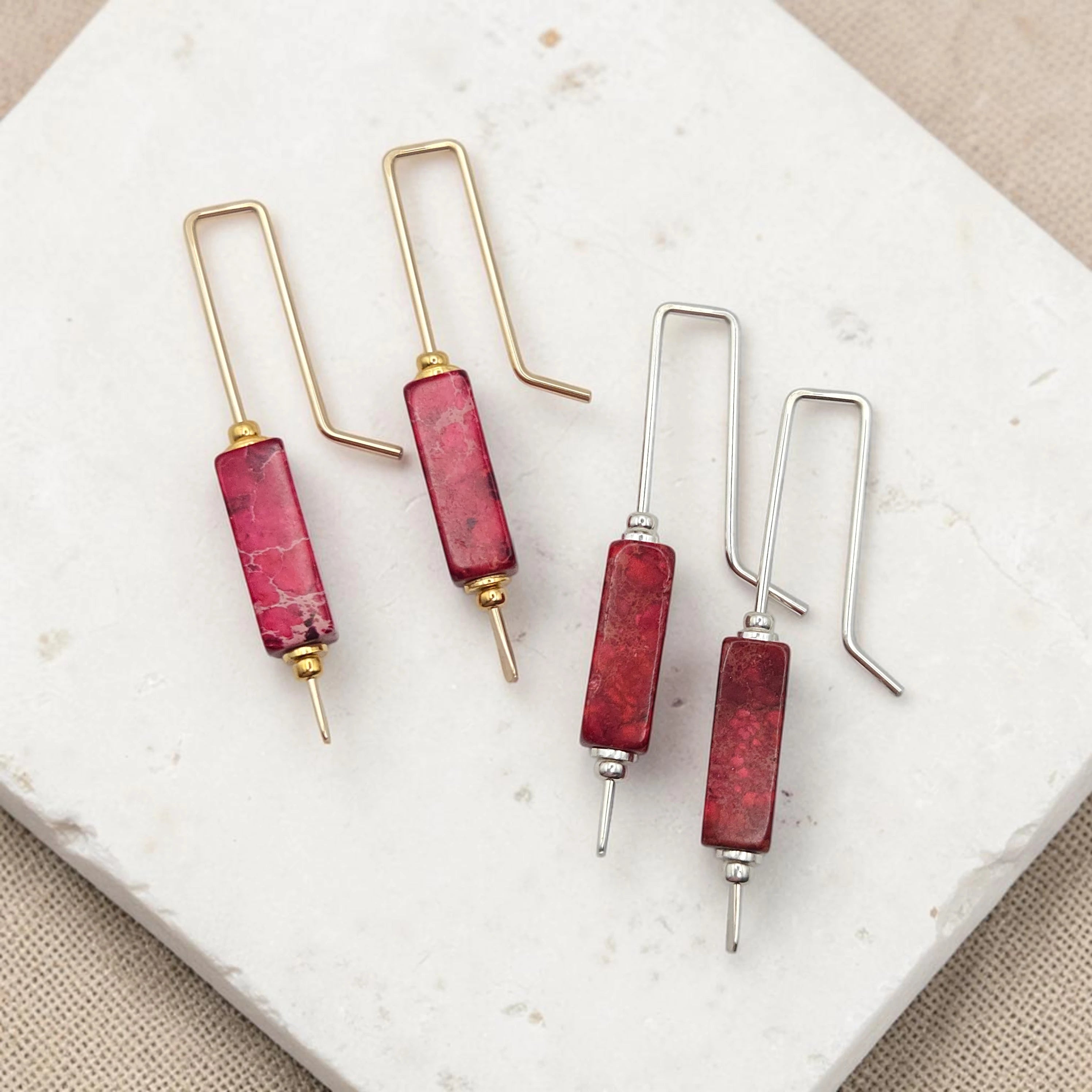 Set of earrings with red cylindrical beads and gold and silver hooks on a marble surface.