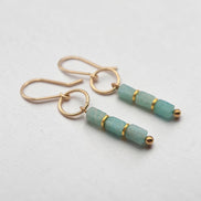 A pair of gold drop earrings featuring three faceted light green Amazonite beads separated by thin gold bands, with small gold beads at the ends, shown on a light gray textured background.