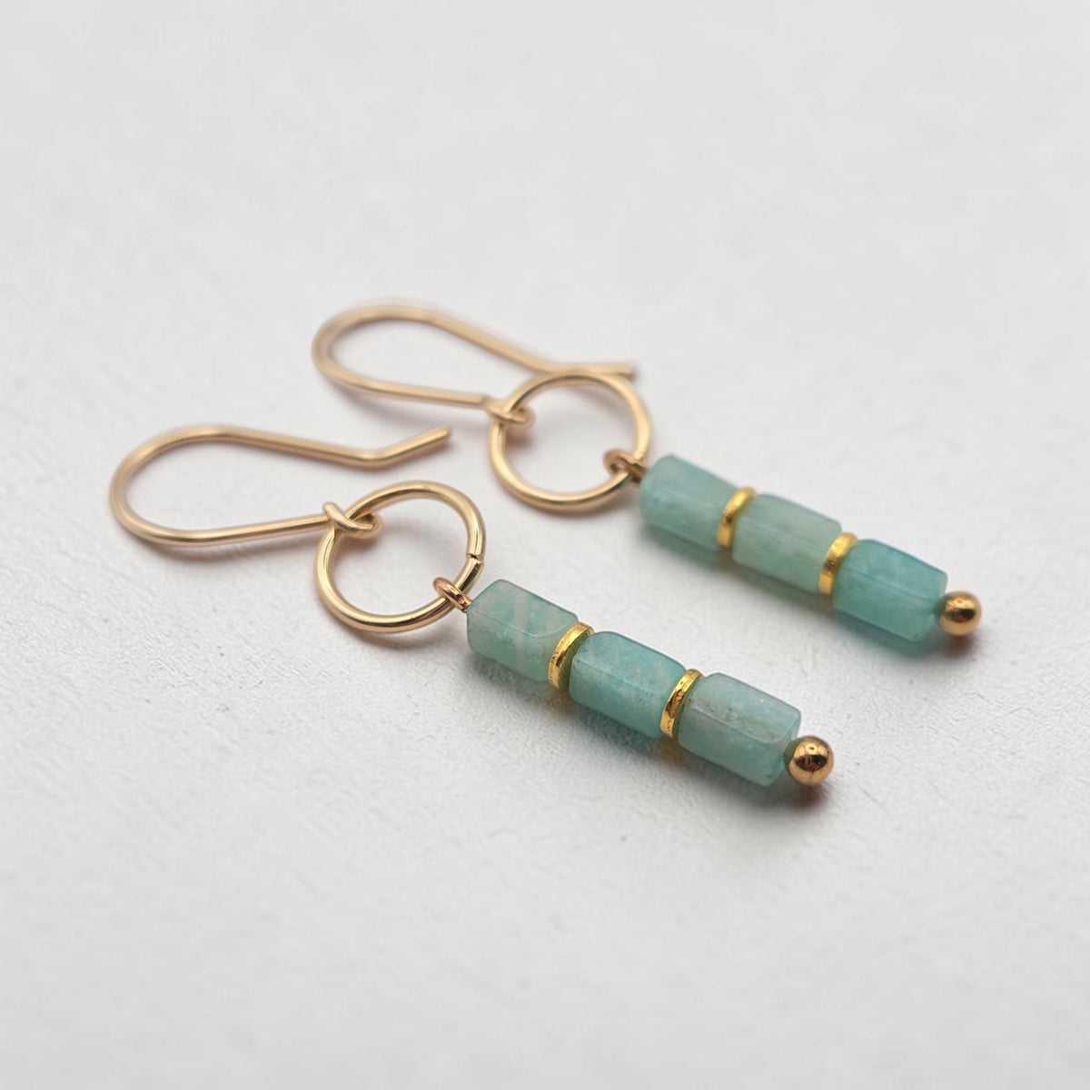 A pair of gold drop earrings featuring three faceted light green Amazonite beads separated by thin gold bands, with small gold beads at the ends, shown on a light gray textured background.