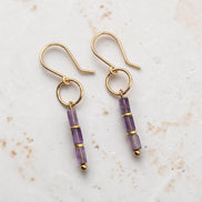 A pair of gold dangle earrings with three faceted amethyst beads on each earring, displayed on a light textured stone surface.