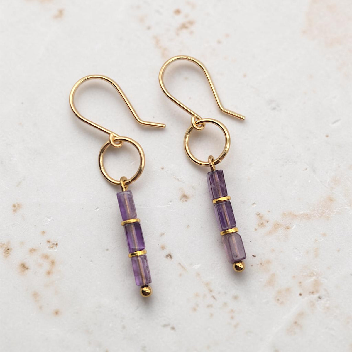 A pair of gold dangle earrings with three faceted amethyst beads on each earring, displayed on a light textured stone surface.