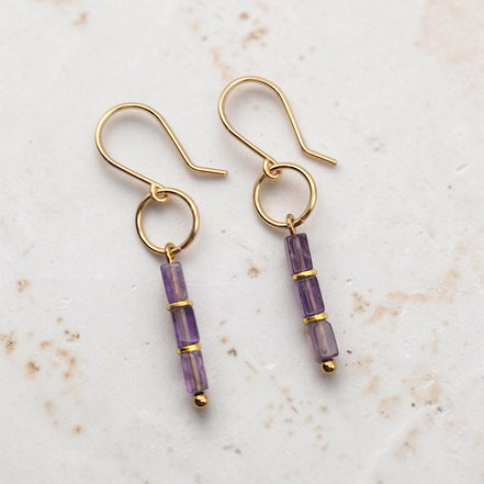 A pair of gold dangle earrings with three faceted amethyst beads on each earring, displayed on a light textured stone surface.