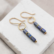 A pair of gold-tone dangle earrings with rectangular lapis lazuli beads, displayed on a white textured stone block.
