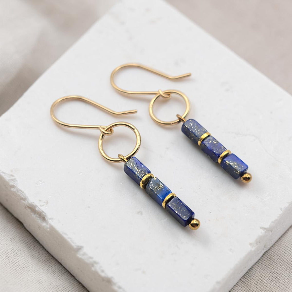 A pair of gold-tone dangle earrings with rectangular lapis lazuli beads, displayed on a white textured stone block.