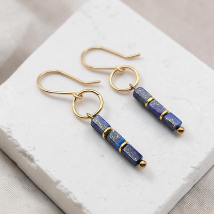 A pair of gold-tone dangle earrings with rectangular lapis lazuli beads, displayed on a white textured stone block.