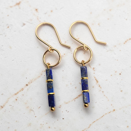 A pair of gold dangle earrings with rectangular lapis lazuli beads, displayed on a light textured marble background.