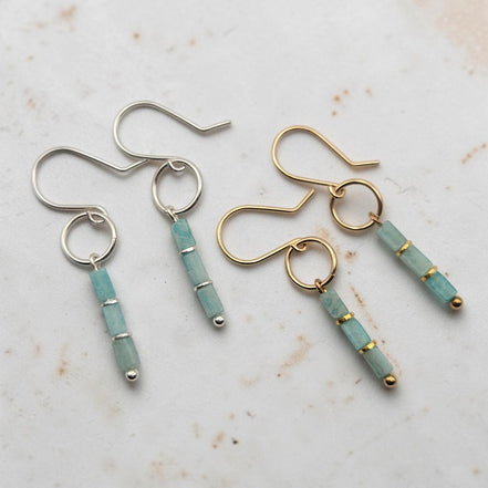 Two pairs of dangle earrings on a light textured surface. One pair is sterling silver with a round hoop and a dangling cylindrical bead cluster in light aqua amazonite stone with small gold spacer rings.