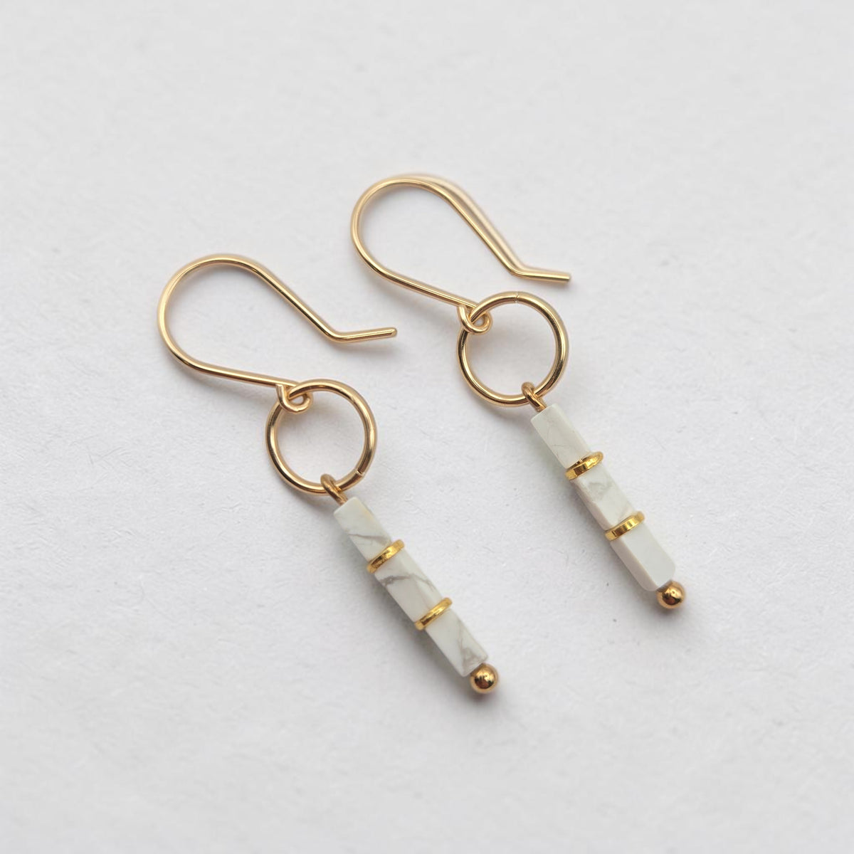 A pair of gold drop earrings with white marble beads, displayed on a clean, light gray textured background.