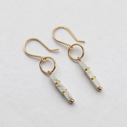 A pair of gold drop earrings with white marble beads, displayed on a clean, light gray textured background.