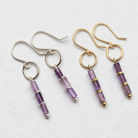 two drop earrings on a light gray background, featuring amethyst faceted beads with silver and gold metal settings and hooks.