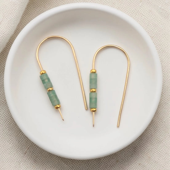 Gold and green beaded earrings on a white dish with a beige fabric background
