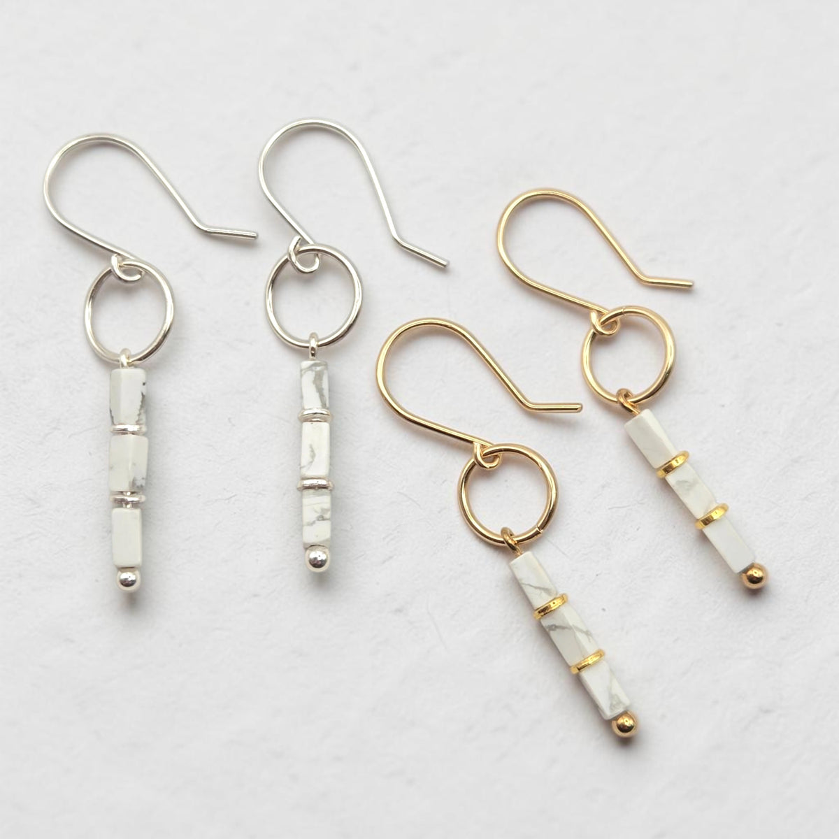 Four dangle earrings arranged on a light textured background. Two earrings have silver-tone hooks and bezel-set white marbled beads, while two have gold-tone hooks and bezel-set white marbled beads with small gold accents.