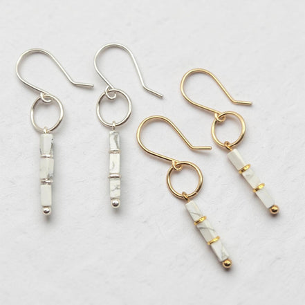 Four dangle earrings arranged on a light textured background. Two earrings have silver-tone hooks and bezel-set white marbled beads, while two have gold-tone hooks and bezel-set white marbled beads with small gold accents.