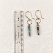 A pair of gold dangle earrings with green and black stone beads, shown next to a stainless steel ruler for scale on a textured off-white background.