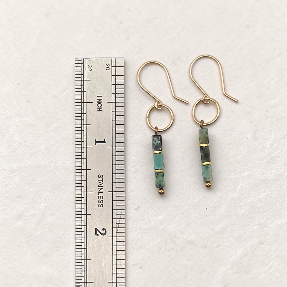 A pair of gold dangle earrings with green and black stone beads, shown next to a stainless steel ruler for scale on a textured off-white background.