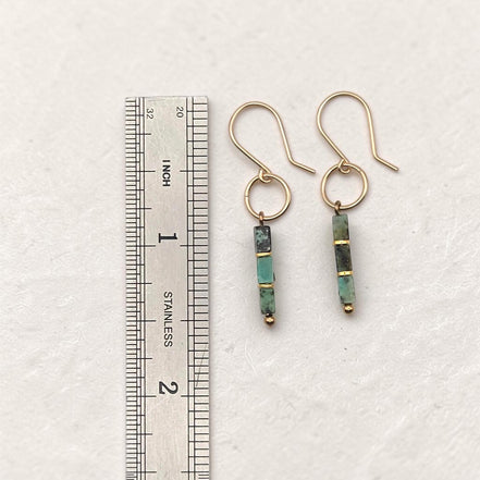 A pair of gold dangle earrings with green and black stone beads, shown next to a stainless steel ruler for scale on a textured off-white background.
