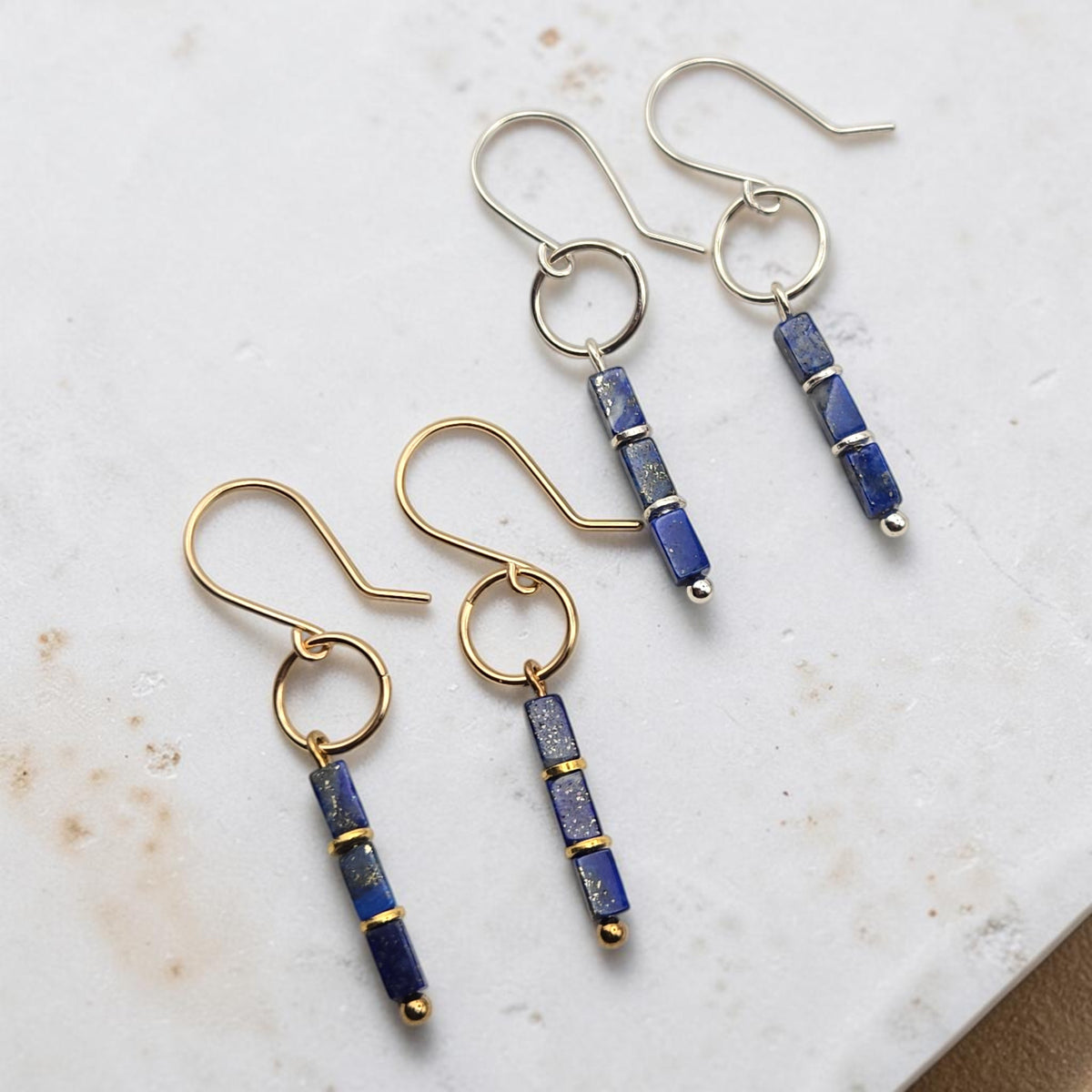 Two pairs of dangle earrings on a light marble surface. Each pair has a gold-tone or silver-tone fishhook ear wire with a small circular ring, connected to a vertical rectangular bead strand made of deep blue lapis lazuli-style beads separated by thin gold-tone bands.