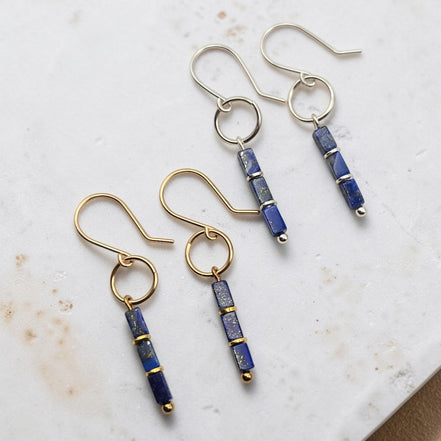 Two pairs of dangle earrings on a light marble surface. Each pair has a gold-tone or silver-tone fishhook ear wire with a small circular ring, connected to a vertical rectangular bead strand made of deep blue lapis lazuli-style beads separated by thin gold-tone bands.