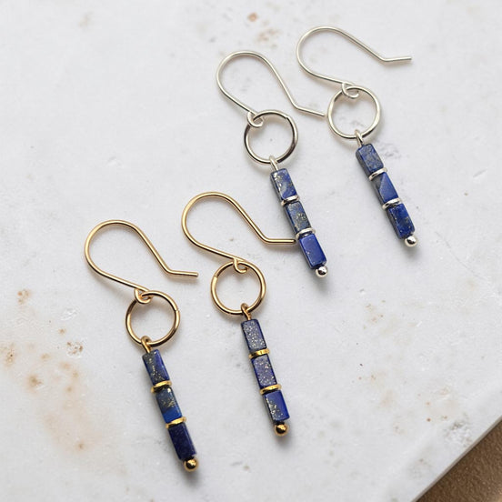 Two pairs of dangle earrings on a light marble surface. Each pair has a gold-tone or silver-tone fishhook ear wire with a small circular ring, connected to a vertical rectangular bead strand made of deep blue lapis lazuli-style beads separated by thin gold-tone bands.
