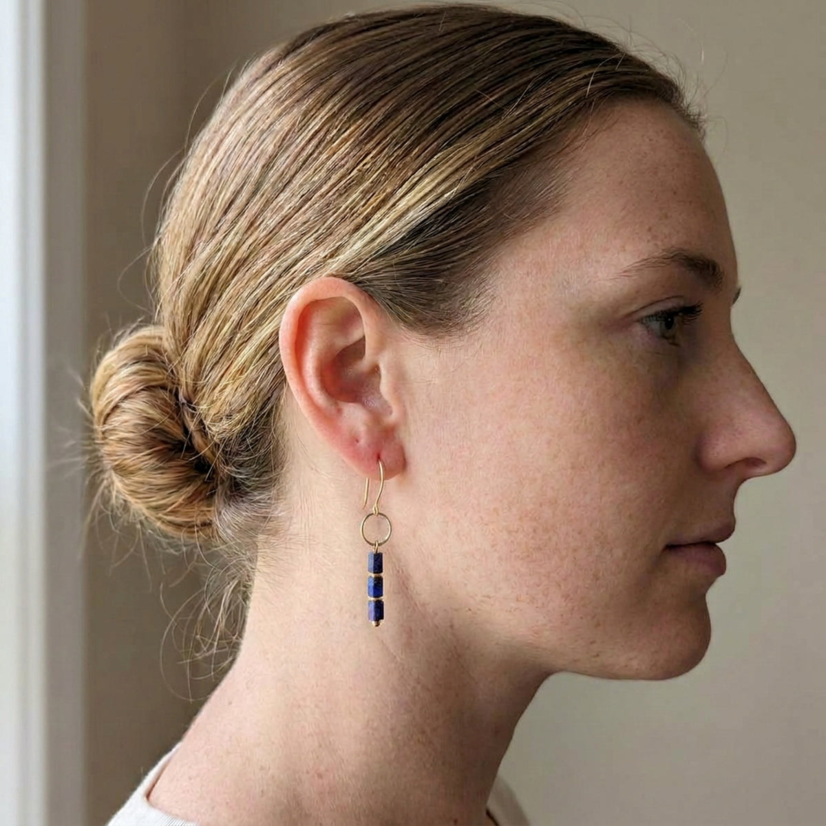A woman in profile wearing a delicate gold dangle earring with three small dark blue beads, shown against a soft beige background.