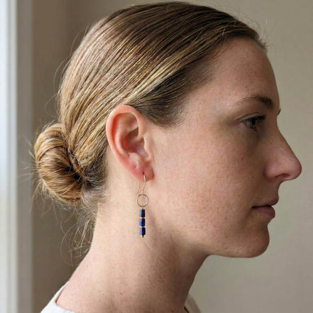 A woman in profile wearing a delicate gold dangle earring with three small dark blue beads, shown against a soft beige background.