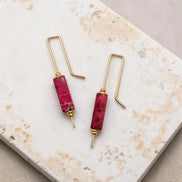 Pair of earrings with red cylindrical beads and gold hooks on a marble surface