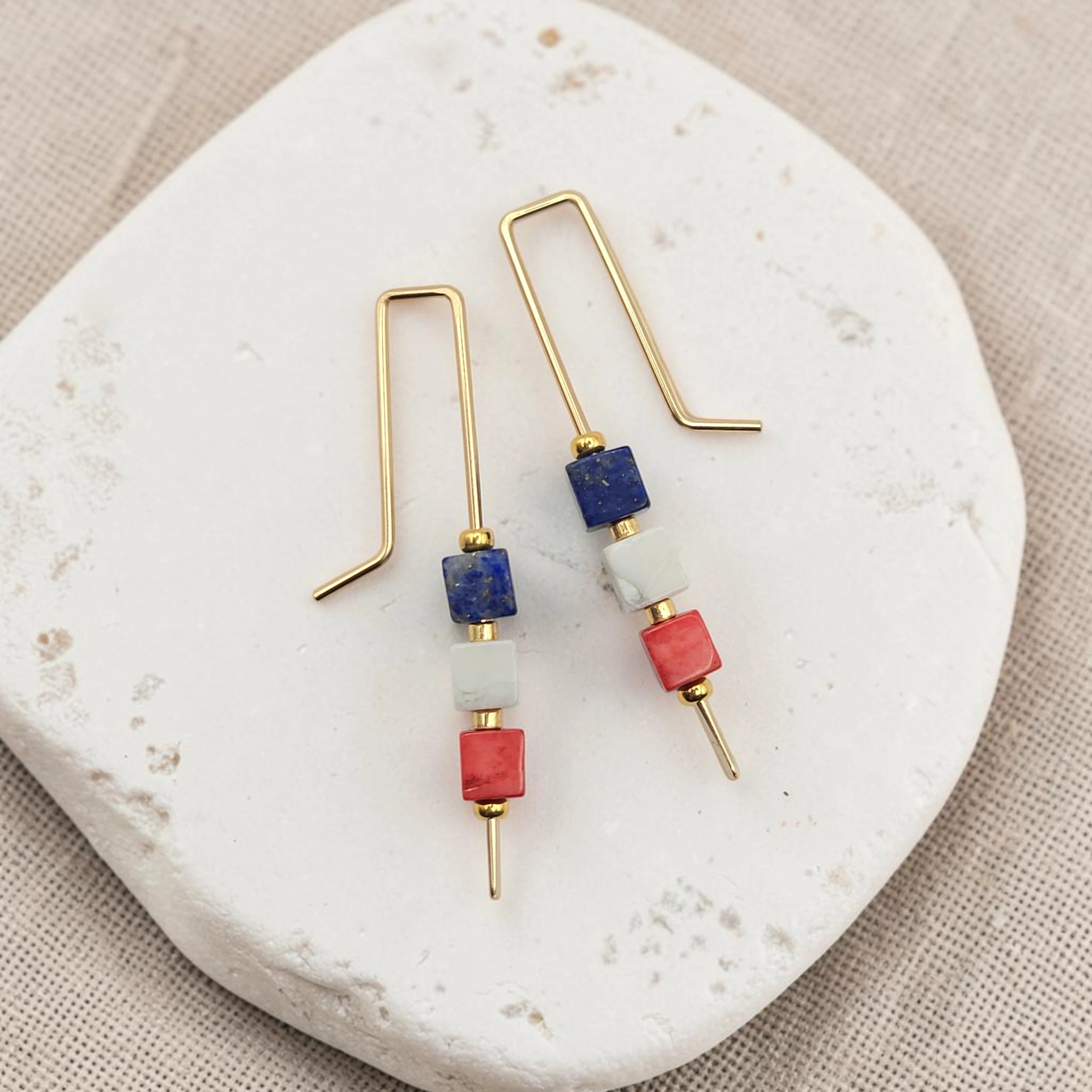 Pair of earrings with gold hooks, blue, and red beads on a white stone surface.