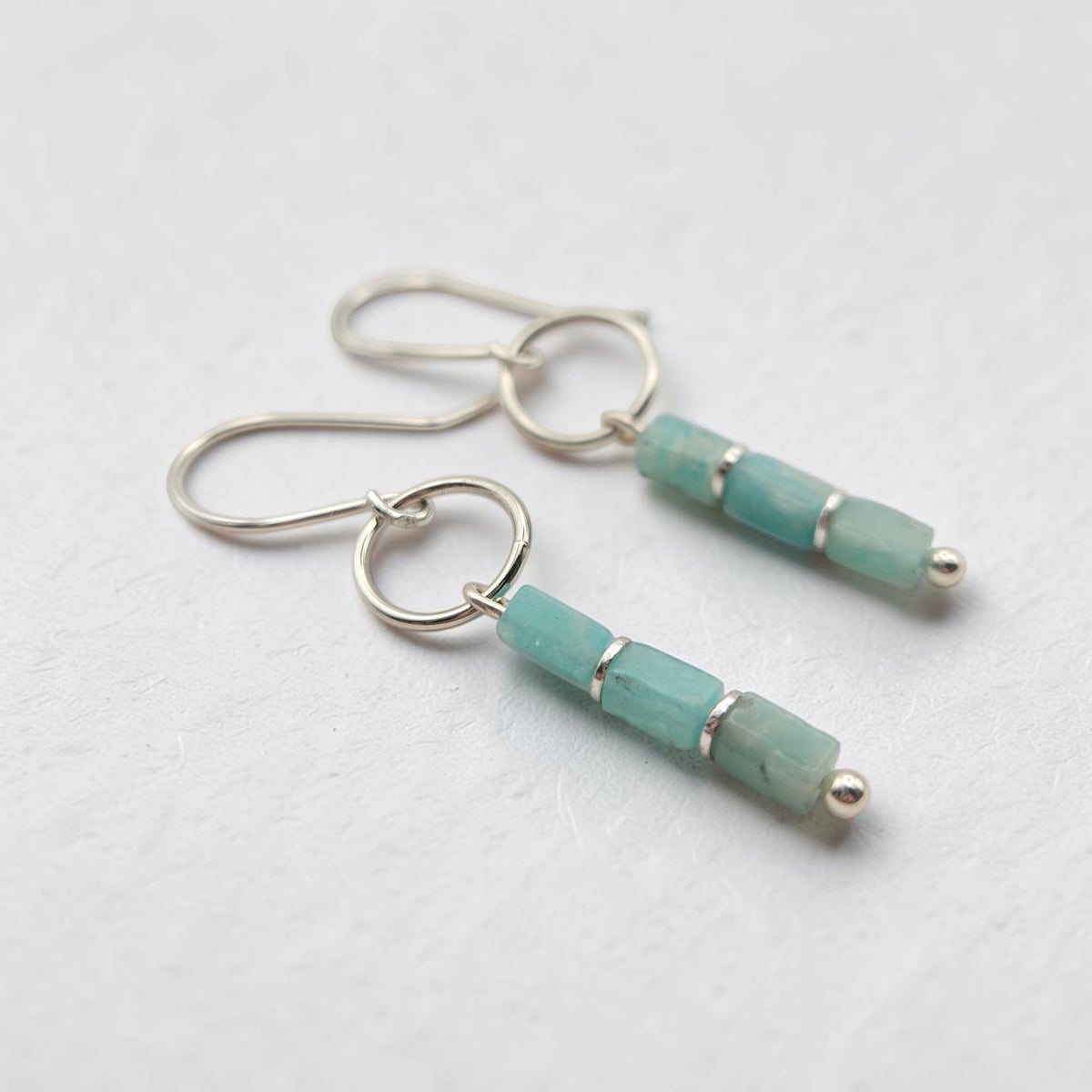 A pair of silver dangle earrings featuring three faceted light blue-green amazonite beads on each earring, displayed on a clean, light gray textured surface.
