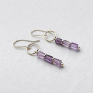 A pair of silver drop earrings with three faceted amethyst beads on each earring, displayed on a textured light gray background.