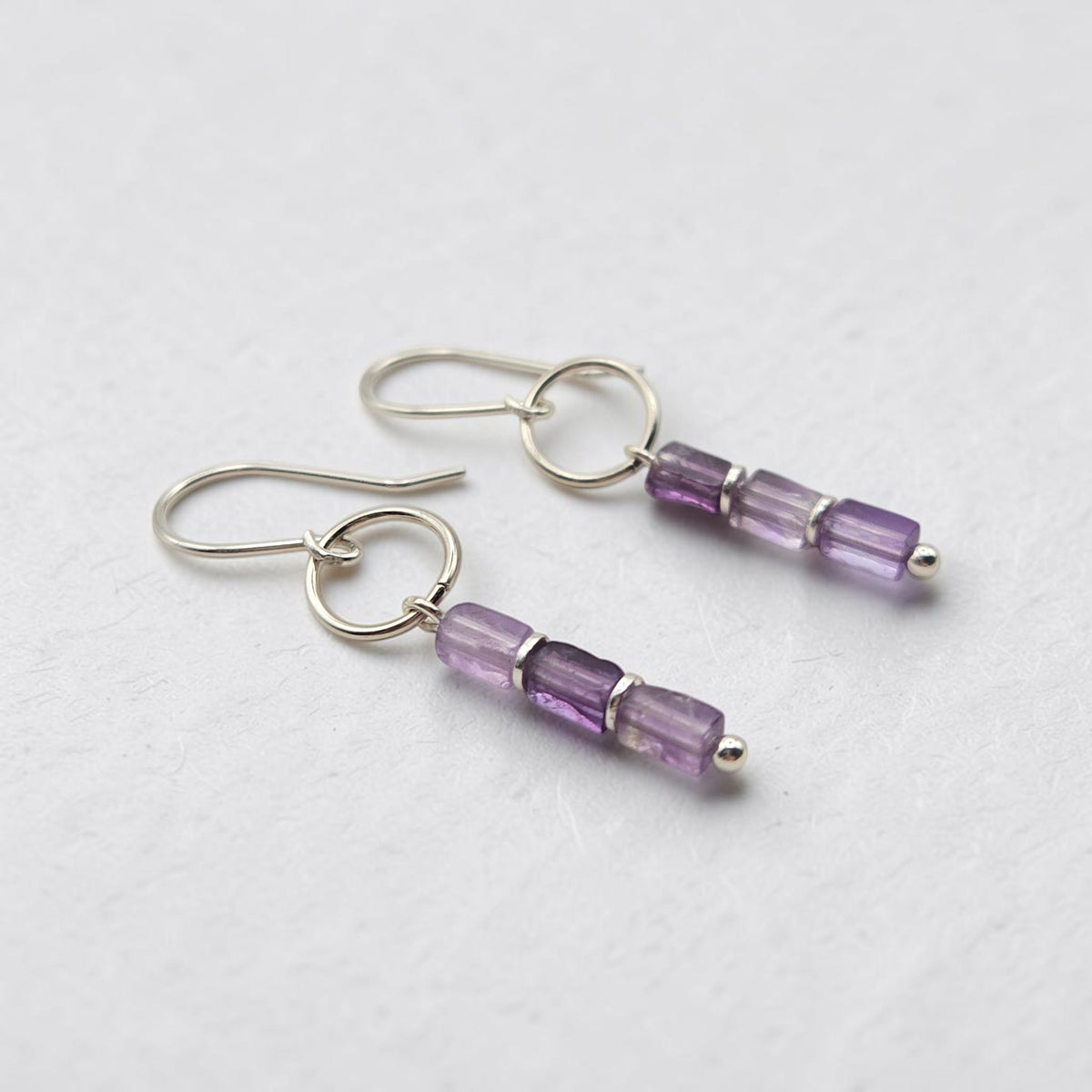A pair of silver drop earrings with three faceted amethyst beads on each earring, displayed on a textured light gray background.