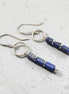 A pair of silver drop earrings featuring rectangular lapis lazuli beads with natural gold flecks, displayed on a textured light gray surface.