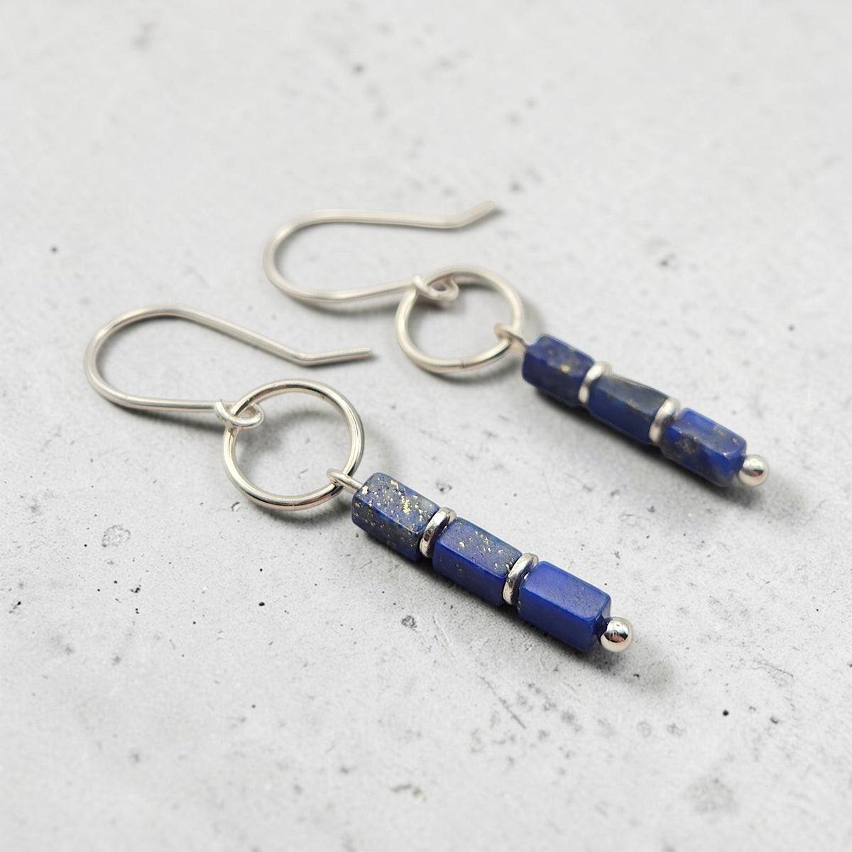 A pair of silver drop earrings featuring rectangular lapis lazuli beads with natural gold flecks, displayed on a textured light gray surface.