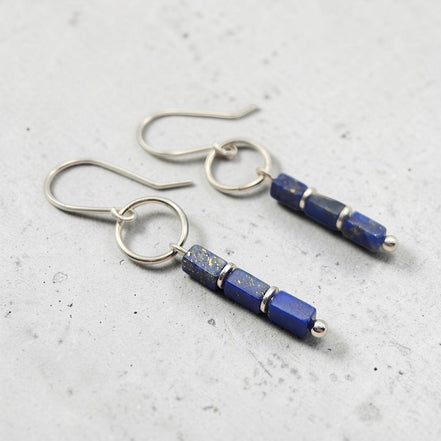 A pair of silver drop earrings featuring rectangular lapis lazuli beads with natural gold flecks, displayed on a textured light gray surface.