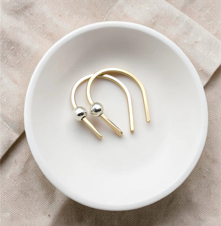 tiny handmade gold huggie earrings with a silver bead laying in a white porcelain bowl