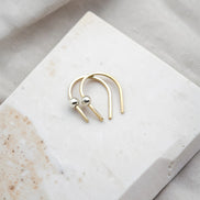 tiny handmade gold huggie earrings with a silver bead laying on a white and tan travertine block