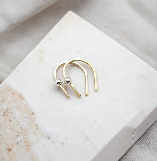 tiny handmade gold huggie earrings with a silver bead laying on a white and tan travertine block